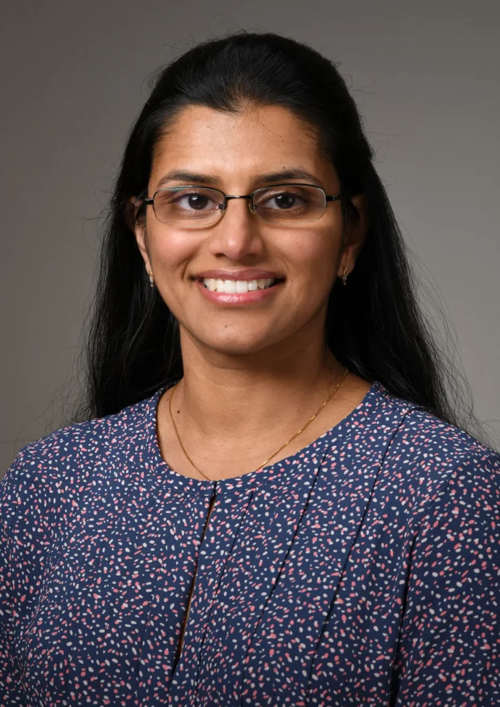 Neethu Menon, MD is pictured wearing glasses, a blue patterned blouse. Dr Neethu Menon is smiling, her hair is pinned back.
