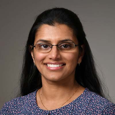 Neethu Menon, MD is pictured wearing glasses, a blue patterned blouse. Dr Neethu Menon is smiling, her hair is pinned back.