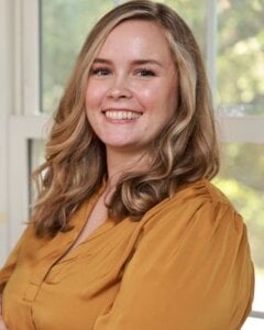 Image shows caucasian woman with long blonde hair, she is smiling, she is wearing a mustard color blouse. The image shows Leah Singer, licensed counsellor.